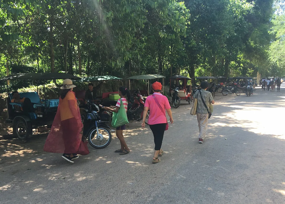 Tuk-tuks parked under trees with visitors walking past, near Ta Prohm in Angkor, seen on Angkor Wat tours from Siem Reap